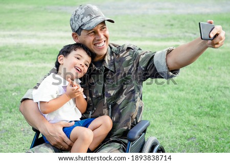 Joyful disabled military dad and his little son taking selfie together in park. Boy sitting on dads lap. Veteran of war or disability concept
