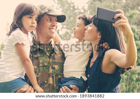 Joyful military man holding and hugging two kids while his wife taking family selfie on cell.. Medium shot. Family reunion or returning home concept