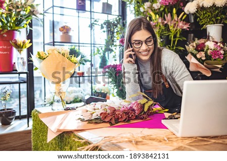 Smiling Woman Florist Small Business Flower Shop Owner. She is using her telephone and laptop to take orders for her store. Female gardener noting client order during mobile phone conversation Royalty-Free Stock Photo #1893842131