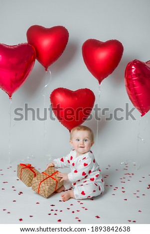 Happy cute baby kid toddler in pajamas sits on the floor strewn with heart-shaped confetti and with presents. Red balloons on the background. St. Valentine's Day concept. Vertical picture