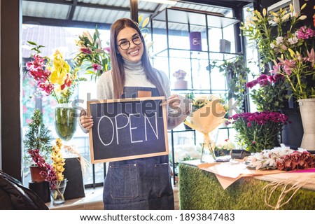Woman florist holds a sign saying open on flower shop hang on door at entrance. After quarantine. Business opening. Young business owner holding OPEN sign near flower shop. Space for text