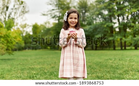 The horizontal image of a joyful little girl dressed in a pink dress smiles, and makes a wish with the birthday cupcake in her hands in the park. A happy cute child celebrates her birthday outdoor.