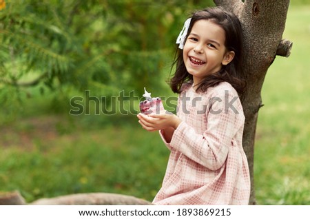 Image of a cheerful little girl smiles and makes a wish with the birthday cupcake in her hands in the park. Happy cute kid celebrates her birthday outdoor.