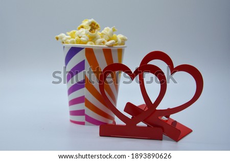 Selective focus of dark red metallic candlesticks in form of hearts and light beige popcorn in bright multicolored disposable paper cup in light of lamps on white background 