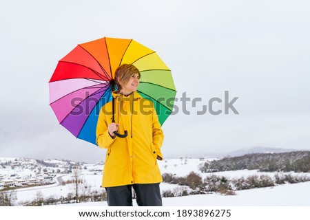 portrait of a happy woman carrying a multicolored umbrella like a rainbow in the snow on a mountain with a village making a beautiful landscape and wearing a yellow jacket making a colorful picture