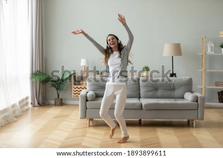 Overjoyed woman wearing headphones dancing in modern living room, having fun, moving to popular music at home, excited attractive young female enjoying leisure time, listening to favorite song Royalty-Free Stock Photo #1893899611
