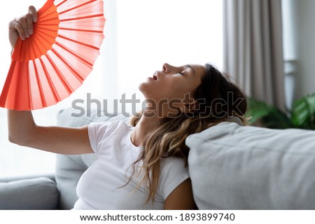 Close up overheated woman waving paper fan, breathing air, leaning back on couch alone, exhausted sick young female feeling unwell, suffering from heating, fever or hot summer weather at home Royalty-Free Stock Photo #1893899740