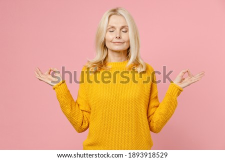 Smiling elderly gray-haired blonde woman lady 40s 50s years old in yellow sweater hold hands in yoga gesture relaxing meditating, trying to calm down isolated on pink color background studio portrait