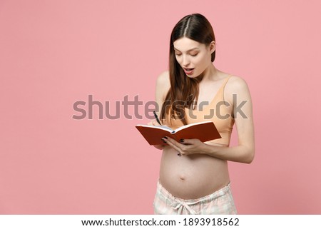 Young calm pregnant woman keeping hands on big belly stomach tummy with baby hold pen writes notes in notebook isolated on pastel pink background studio. Maternity family pregnancy expectation concept