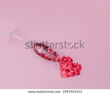 Champagne glass filled with heart shaped confetti and heart made of confetti on the pink background. Celebrating Valentine's day.