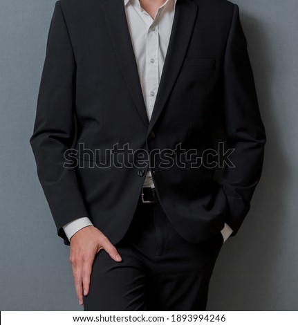 A male in a classic black suit with his hand in the pocket leaning on a wall