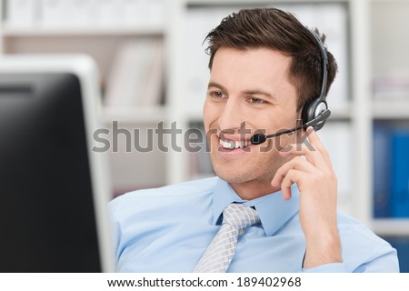 Smiling friendly handsome young male call centre operator or client services personnel beaming as he listens to a call and checks information on his computer monitor Royalty-Free Stock Photo #189402968
