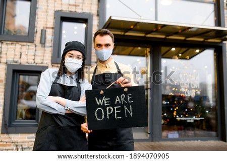 Welcome, we are OPEN. Two friendly waiters wearing protective masks. Multiracial waiters, caucasian guy and asian girl standing outside a restaurant, cafe or bar and showing signboard OPEN