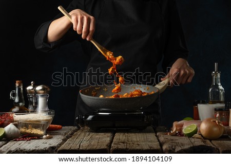 Chef in black uniform mix frying chicken fillet with sauce. Backstage of cooking traditional Indian chicken curry on dark blue background. Concept cooking process. Hot recipe. Frozen motion.