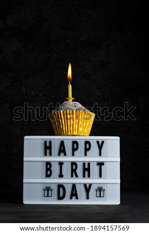 Blackboard with happy birthday lettering and cupcake in paper cup with yellow candle with flame in low key greeting card