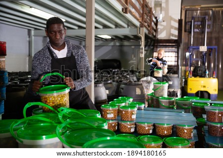 Afro-american male worker stocks plastic containers and cans with olives in warehouse. High quality photo