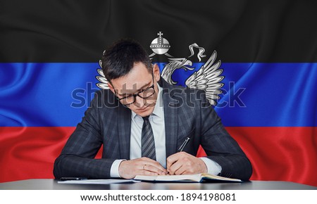 A businessman in a jacket and glasses sits at a table signs a contract against the background of a flag dnr