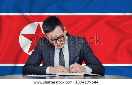 A businessman in a jacket and glasses sits at a table signs a contract against the background of a flag North Korea
