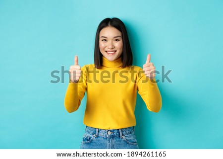 Cheerful asian female model showing thumbs up gesture, smiling and looking impressed, praise good product, standing over blue background Royalty-Free Stock Photo #1894261165