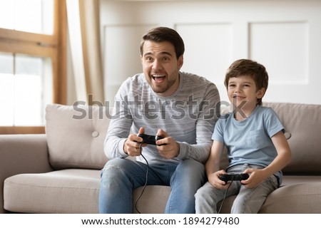 Happy young Caucasian father and little 6s son sit on couch at home have fun playing computer video game together. Overjoyed dad and small boy child enjoy family weekend engaged in funny activity. Royalty-Free Stock Photo #1894279480