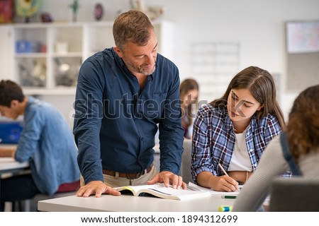 Secondary student being helped by professor during class. Mature man lecturer helping young woman during class test. High school girl in a lecture asking for explanations to helpful teacher.  Royalty-Free Stock Photo #1894312699