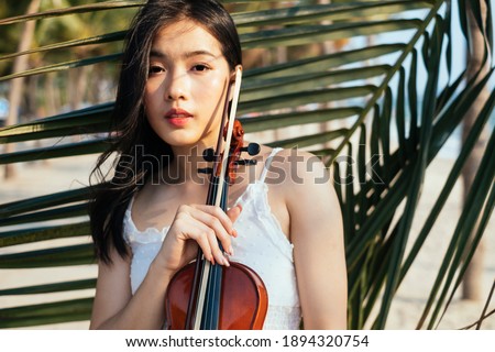 Black long hair violin player woman in white dress and violin standing at the coconut leaf.