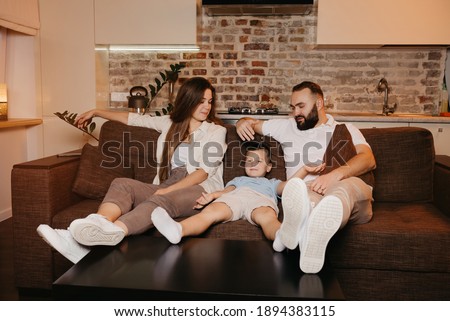 Dad, son, and mom are watching boring TV programs on the sofa in the apartment. The parents are talking to their boy in the evening. Relatives at home. A young mother with long hair is smiling.