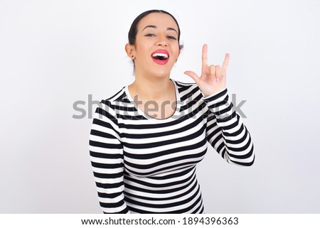 Young beautiful woman wearing stripped t-shirt against white background doing a rock gesture and smiling to the camera. Ready to go to her favorite band concert.
