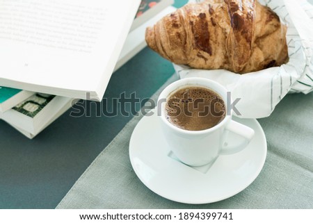 Morning coffee concept, white cup of coffee, empty book page, fresh baked croussant in paper pack on gray cloth and dark grey background