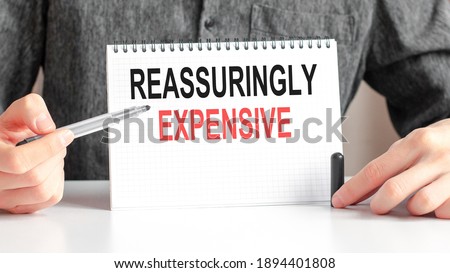 A man in a white holds a piece of paper with the text: REASSURINGLY EXPENSIVE. Multicolored markers and tablet on a table. Business and educational concept for companies and educational institutions.
