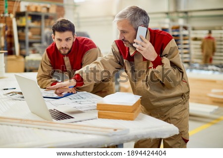 Mid adult worker communicating over mobile phone while using laptop with his colleagues at carpentry workshop.  Royalty-Free Stock Photo #1894424404