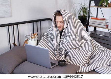 Young attractive caucasian man sitting on a bed wrapped with blanked at home working using laptop with cup of hot drink. Guy browsing Internet for flu, influenza, cold and covid-19 symptoms Royalty-Free Stock Photo #1894443517