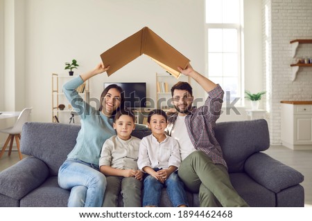 Happy beautiful family of four sitting on couch at home together. Smiling mommy and daddy holding symbolic roof above little kids on sofa. Mortgage, house insurance, future plans, protecting children Royalty-Free Stock Photo #1894462468