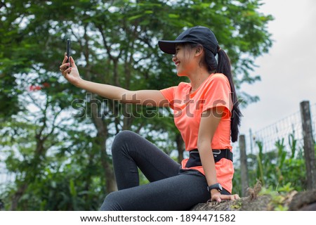 Photo of cheerful young asian sports woman sitting outdoors using mobile phone.
