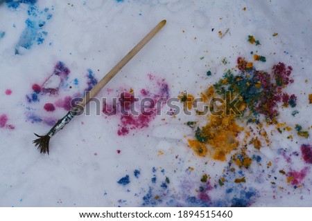 Multi-colored bright splashes of paints on the white snow. The brush lies in the snow. Concept of creativity and hobby.