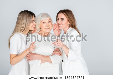 happy woman whispering in ear of senior mother near young daughter isolated on grey