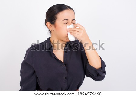 Young ill beautiful Arab woman wearing gray dress against white studio background sneezing her nose on a paper tissue feeling sick. Woman suffering from flu symptoms. 