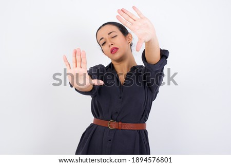 Portrait of smiling young beautiful Arab woman wearing gray dress against white studio background looking at camera and gesturing finger frame. Creativity and photography concept.