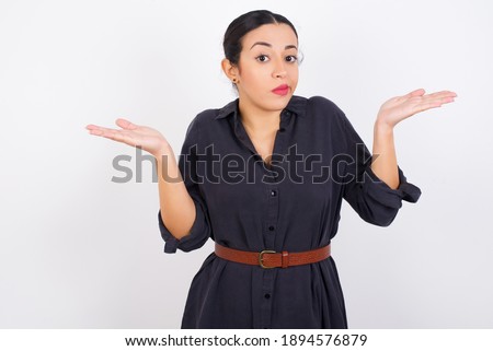 Maybe yes or no. Doubtful young beautiful Arab woman wearing gray dress against white studio background shrugs shoulders in bewilderment, tries to make decision puzzled what he wants.  Royalty-Free Stock Photo #1894576879