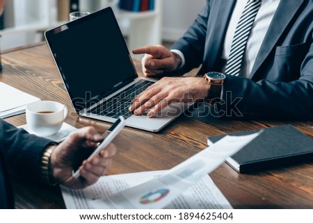 Cropped view of businessman pointing at laptop with blank screen near investor with smartphone and documents on blurred foreground