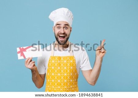 Excited bearded male chef or cook baker man in apron white t-shirt toque chefs hat isolated on blue background. Cooking food concept. Mock up copy space. Hold gift certificate point index finger up