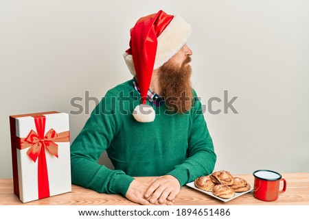 Young irish redhead man wearing christmas sitting on the table with gift looking to side, relax profile pose with natural face with confident smile. 