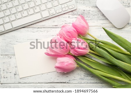 Overhead photo of bouquet of pink tulips lying on wooden desktop with white keyboard computer mouse and paper card with copy space