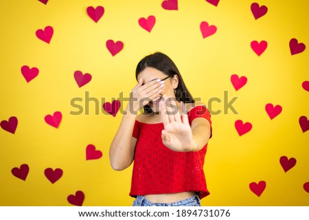 Young beautiful woman over yellow background with red hearts covering eyes with hands and doing stop gesture with sad and fear expression. Embarrassed and negative concept.