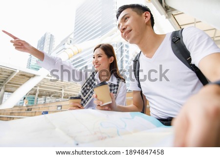 Portrait of cute smiling adult Asian couple tourists sitting on stair while holding paper metro map with woman pointing the direction with blurred tall building background. Selective focus at woman