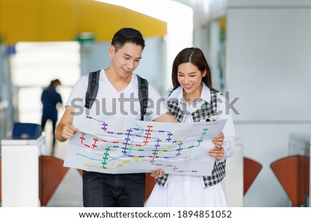 Front view portrait of cute smiling young adult Asian lover travelers standing and looking paper metro map on vacations finding ways to tourist landmarks with blurred Skytrain station background