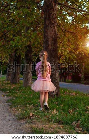 Cheerful little girl with long blonde hair in pink tulle skirt walking alone on empty street on sunset. Happy dreamy cute child on nature background back view. Kids dress fashion. Greeting card.