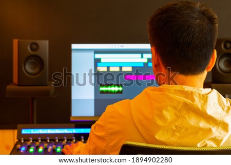 back of asian professional male producer, sound engineer mixing sound on control surface mixer for recording or live broadcasting in studio. post production, recording, broadcasting concept