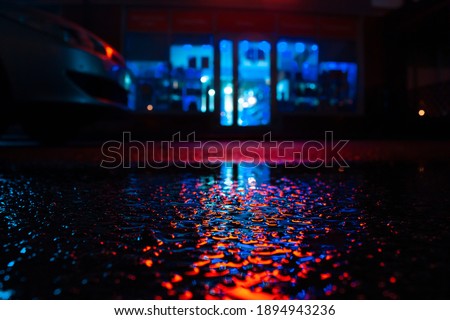 Horizontal night photography of wet with puddle asphalt in focus on the foreground with reflection of illuminated orange sign and blue storefront of a blurred shop on the background in the dark