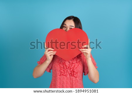 Close-up dark-haired naughty, playful Asian woman in red dress peeps from behind a large red paper heart isolated on a blue background.